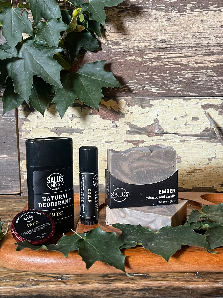 A flat-lay product arrangement featuring a bar of SALUS soap, a black cologne tube, shower bomb, and a black deodorant stick,  placed on a rustic distressed wooden surface with decorative green leaves.