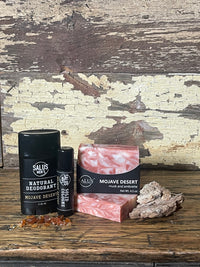 A flat-lay product arrangement of Mojave Desert scented products featuring a bar of SALUS soap, a black cologne tube, shower bomb, and a black deodorant stick,  placed on a rustic distressed wooden surface.