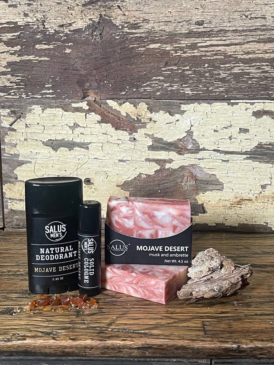 A flat-lay product arrangement of Mojave Desert scented products featuring a bar of SALUS soap, a black cologne tube, shower bomb, and a black deodorant stick,  placed on a rustic distressed wooden surface.