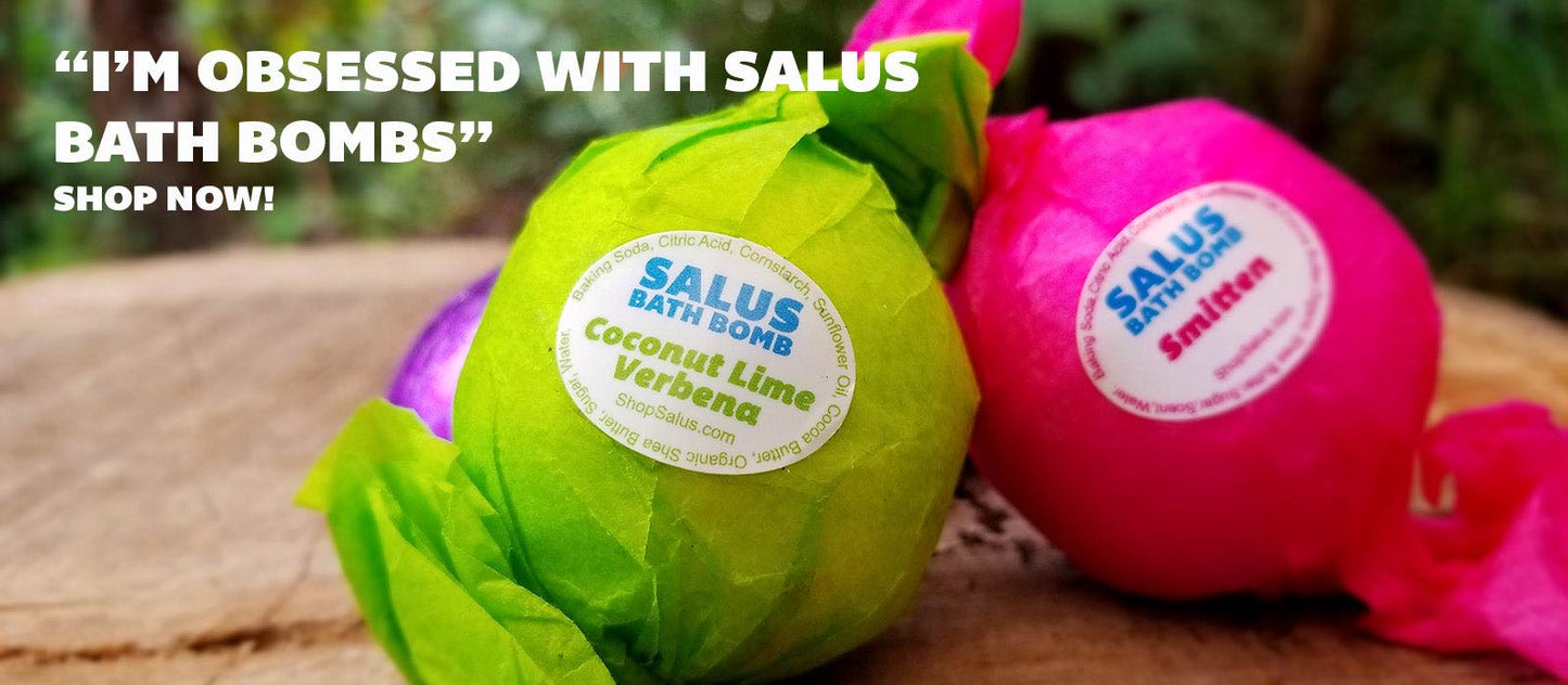 Two SALUS wrapped bath bombs on a wooden surface with an outdoor background. One bath bomb is wrapped in bright green paper and labeled 'Salus Bath Bombs Coconut Lime Verbena,' and the other is wrapped in bright pink paper and labeled 'Smitten'.
