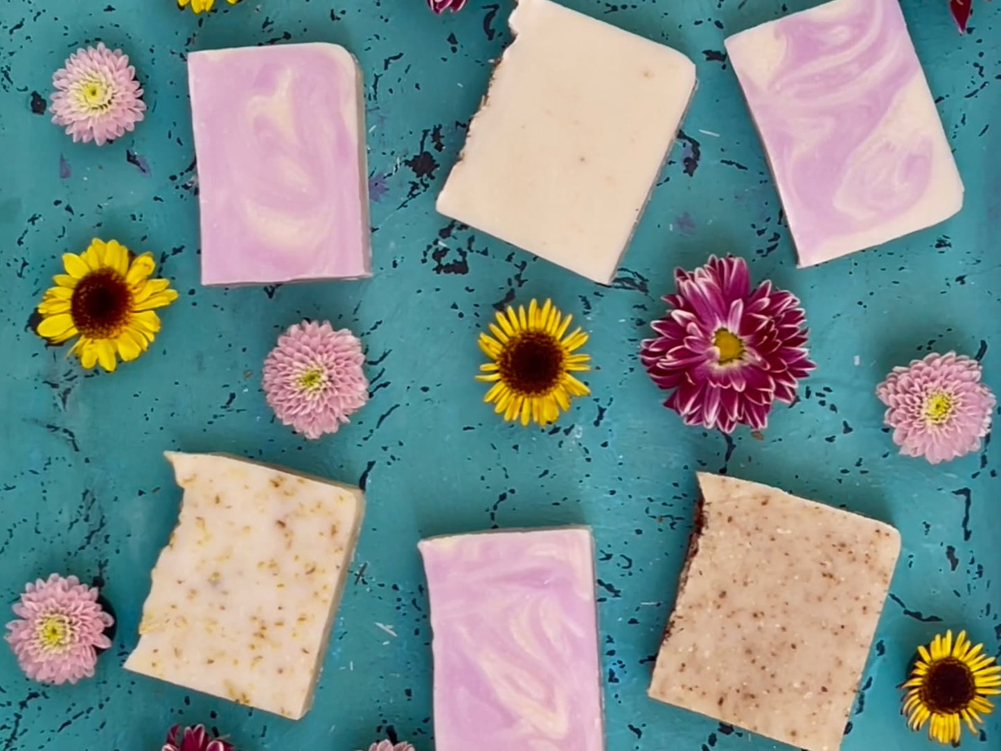 Four rectangular bars of SALUS handmade soap in pastel pink, lavender, cream, and speckled beige tones, arranged on a textured turquoise background with small yellow and pink flowers.