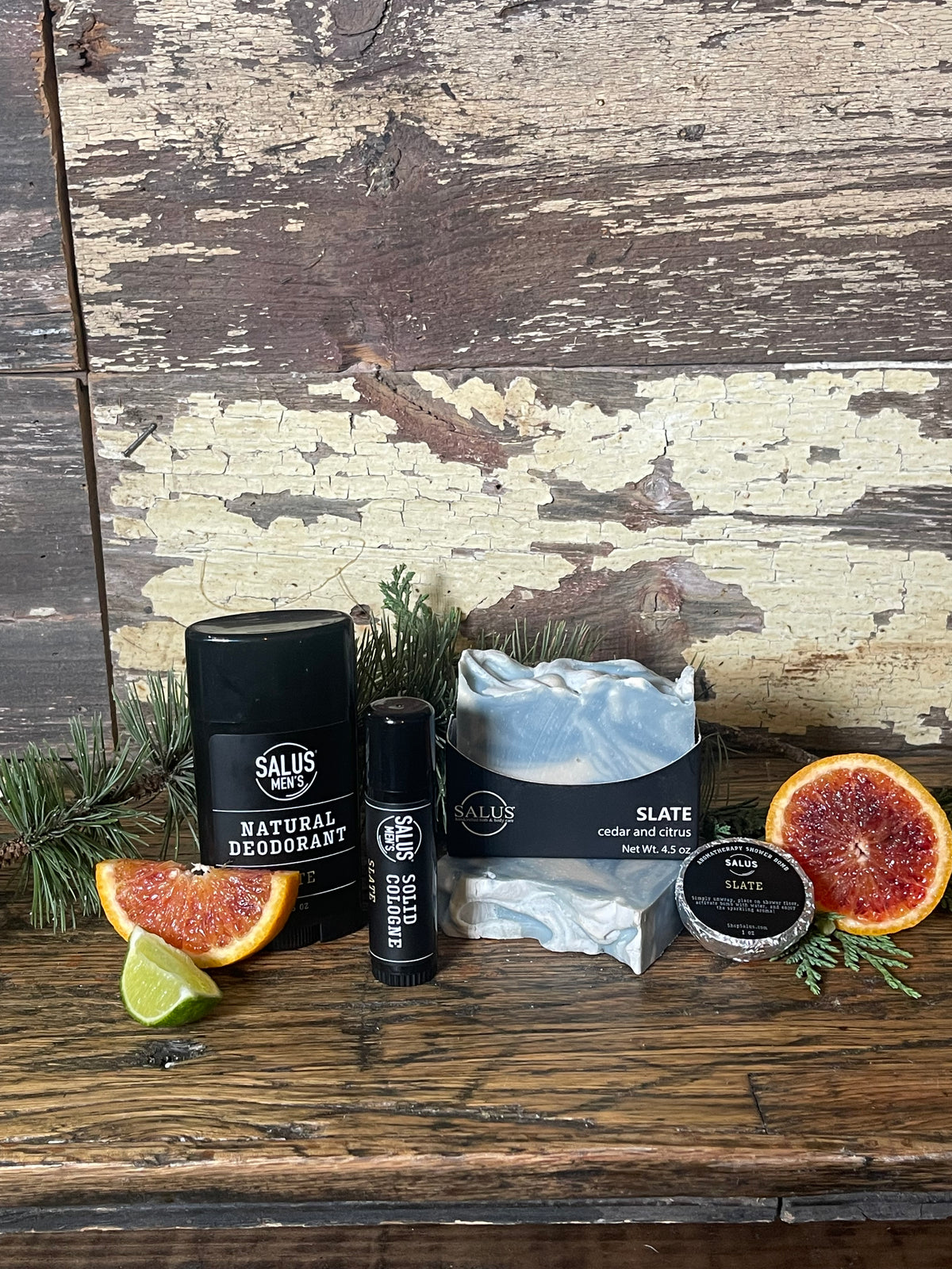A flat-lay product arrangement featuring a bar of SALUS soap, a black cologne tube, shower bomb, and a black deodorant stick,  placed on a rustic distressed wooden surface with decorative green leaves.
