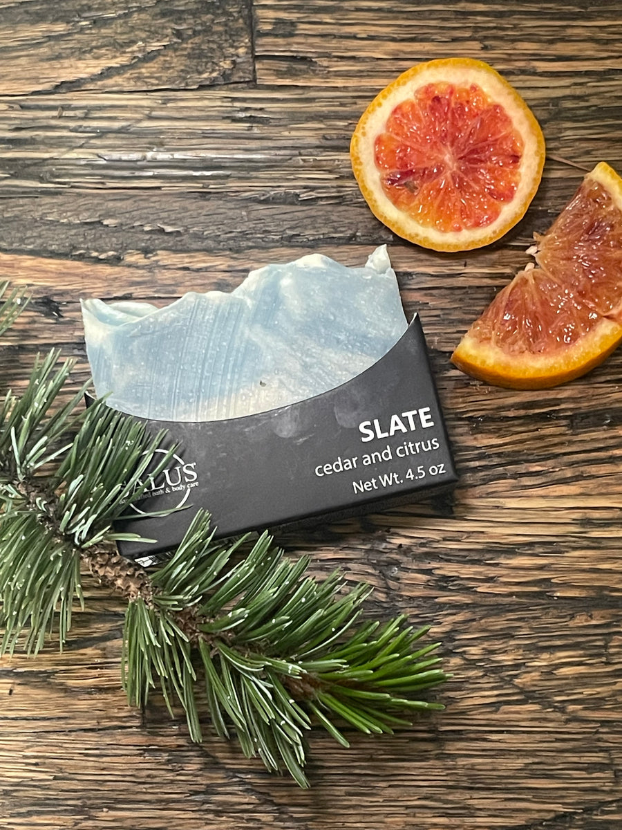 A packaged bar of SALUS soap labeled SLATE cedar and citrus sits on a rustic wooden surface with grapefruit slices and a green pine branch nearby.