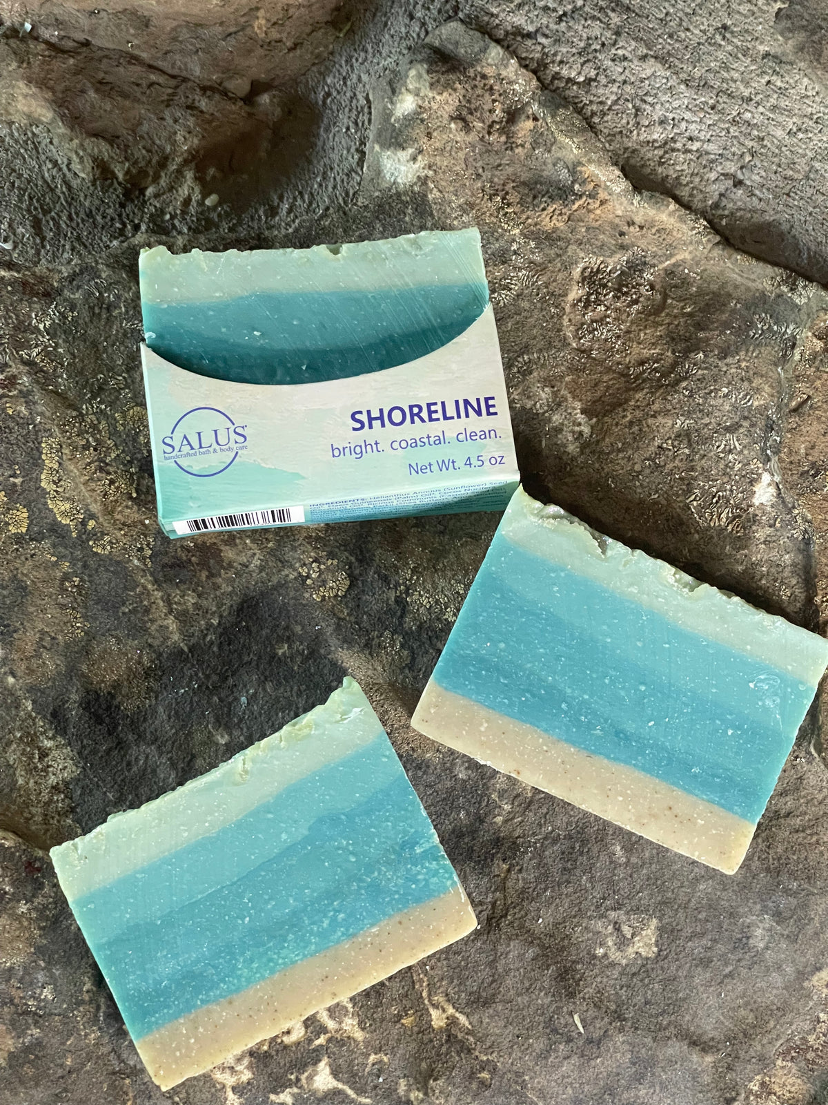 Three bars of SALUS Shoreline soap resting on a textured dark gray stone surface. One bar is upright, showing its white paper label with blue text, while the other two are laid flat, displaying their blue and beige marbled soap layers.